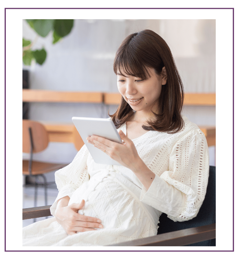 Smiling pregnant woman in a white dress holding her belly and reading from a tablet