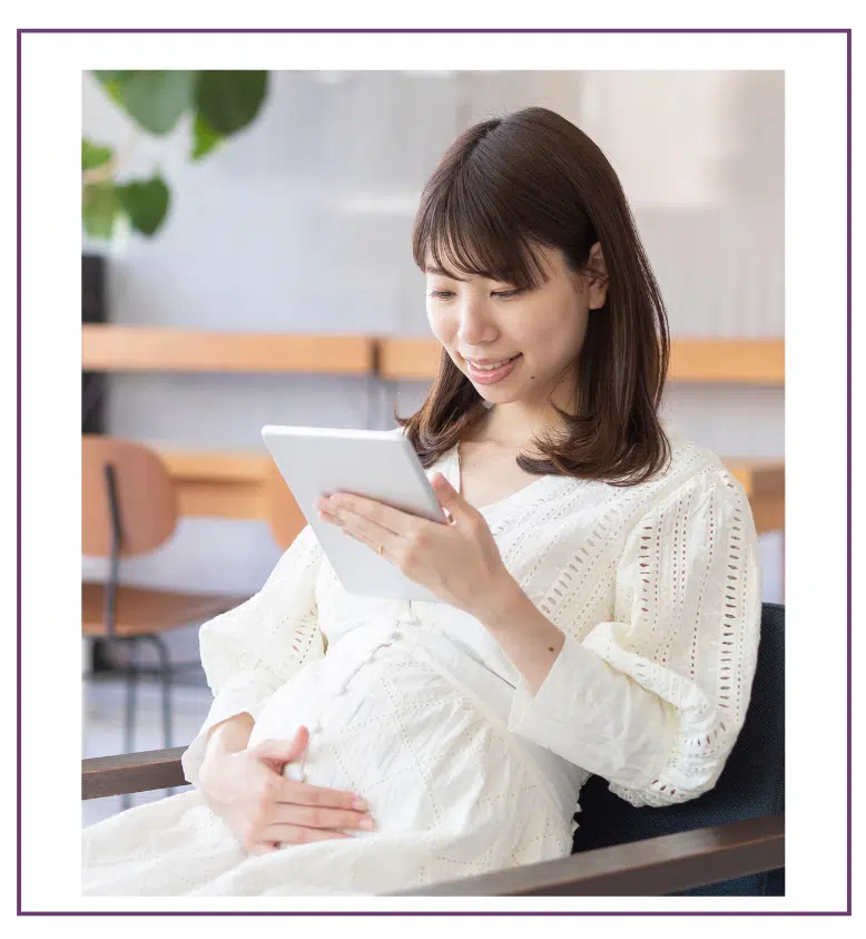 Smiling pregnant woman in a white dress holding her belly and reading from a tablet