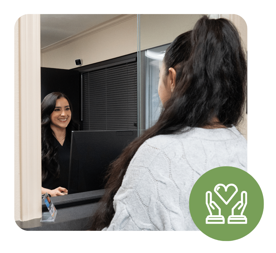 Smiling front desk receptionist greeting a patient in a clinic lobby at the check-in window.